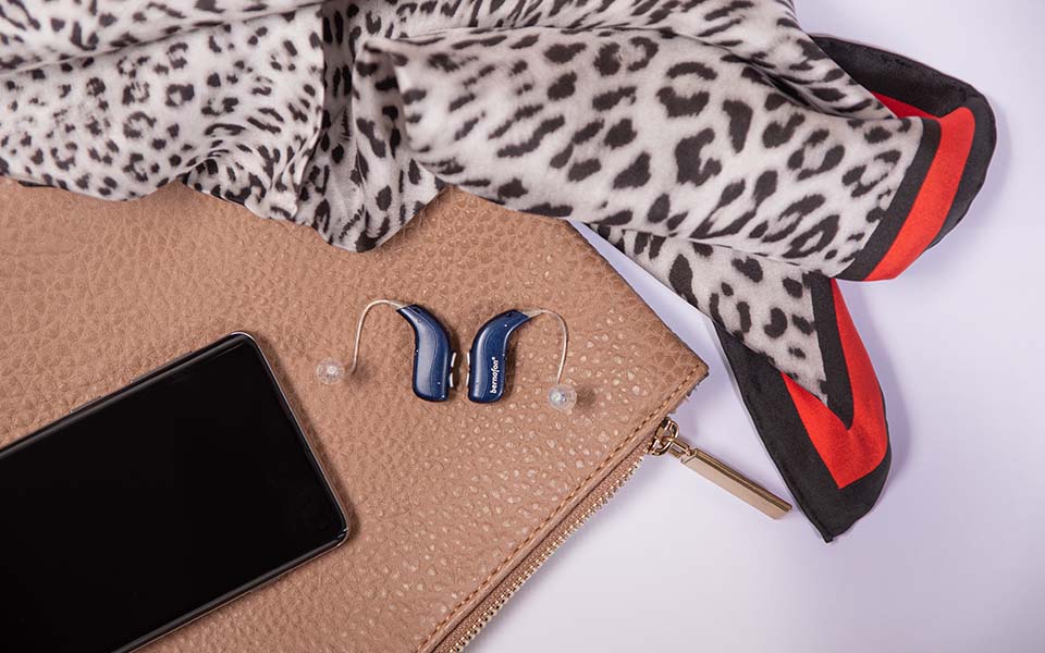 Des appareils auditifs rechargeables Bernafon Alpha bleu nuit sont posés sur un sac à main à côté d'un smartphone et d'un foulard à imprimé animal.