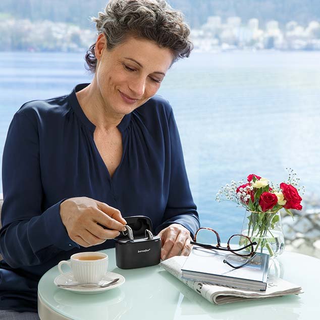 Femme assise à une table avec vue sur un lac suisse et retire ses aides auditives rechargeables Bernafon Alpha de son chargeur portable Charger Plus.