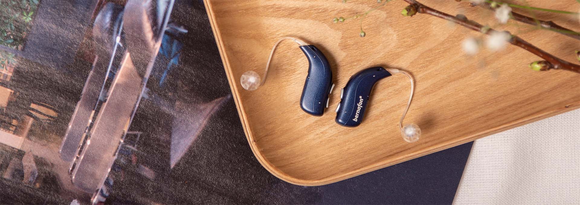 Des appareils auditifs rechargeables Bernafon Alpha bleu nuit placés à côté d'un smartphone sur un chapeau de paille avec un foulard imprimé animal