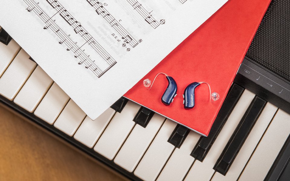 Midnight blue Bernafon Alpha rechargeable hearing aids sit on red notebook on a piano next to a sheet of music notes